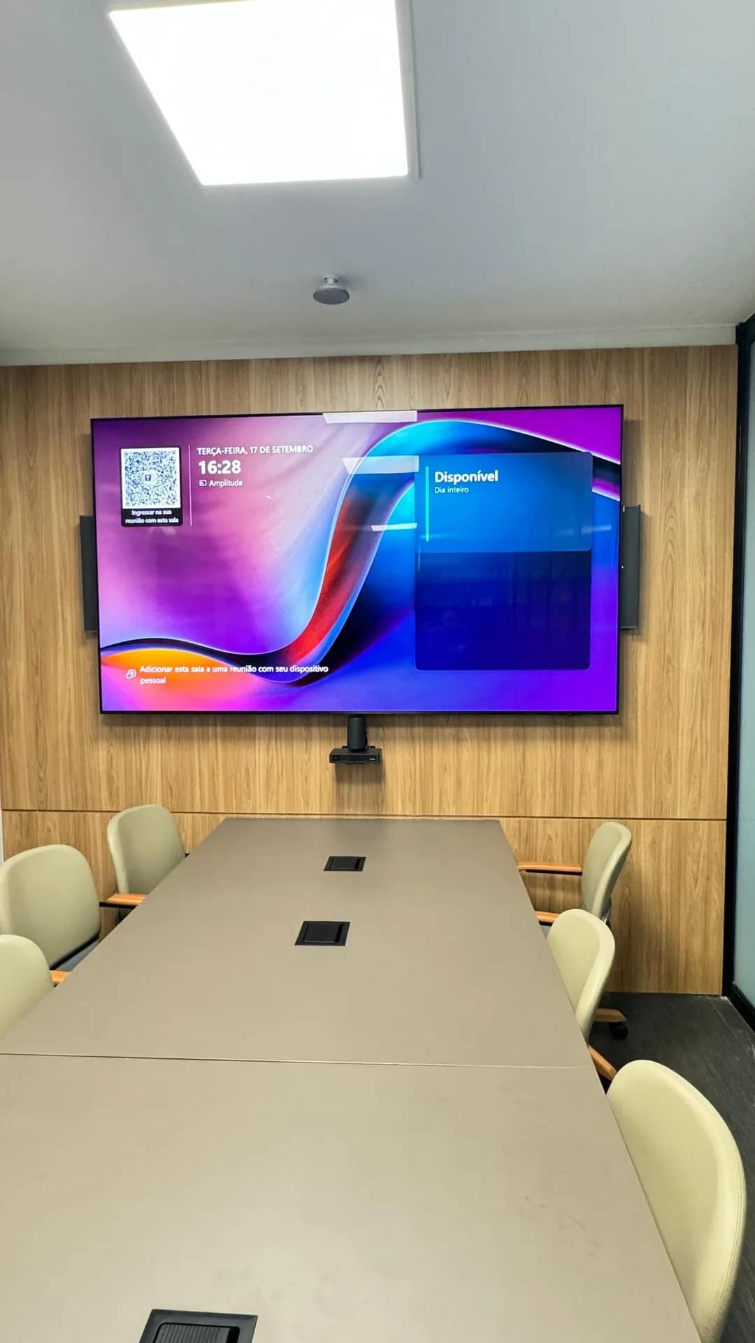 Sala de reunião pronta, com parede de madeira, monitor de videoconferência e mesa com tomadas embutidas.