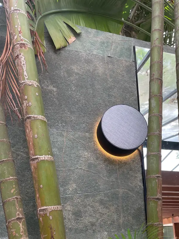 Caixa acústica redonda com luz de contorno, fixada em parede de pedra entre bambus, na Mostra Magna Audio.