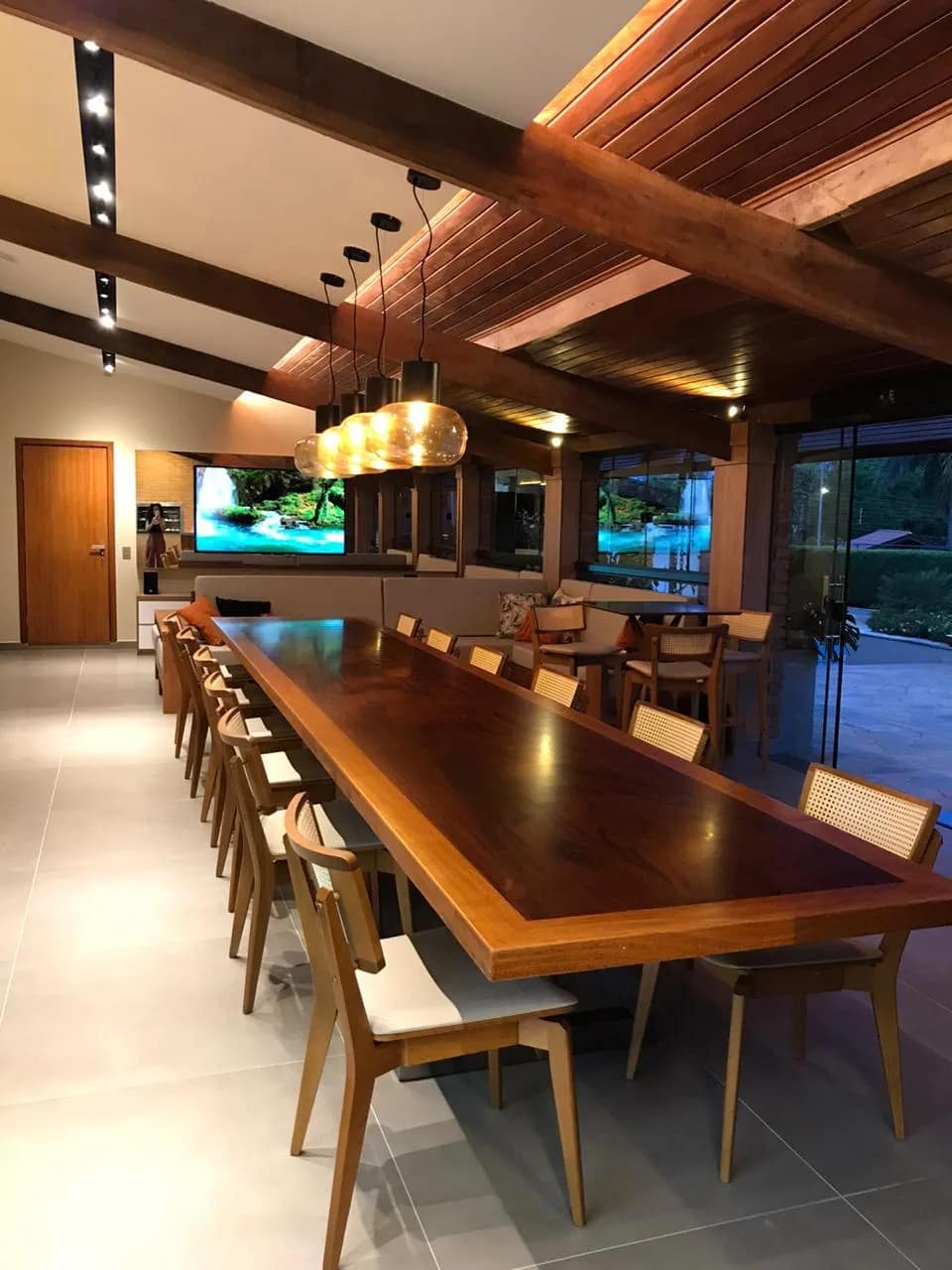 Área gourmet com mesa longa de madeira, pendentes e monitor ao fundo.