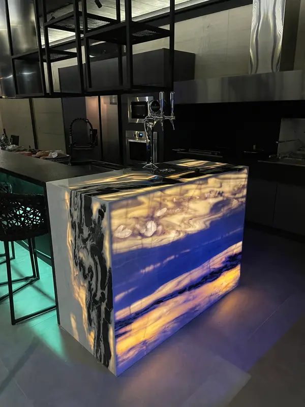 Bancada de ônix iluminada por dentro no espaço gourmet, com banquetas e cozinha ao fundo.