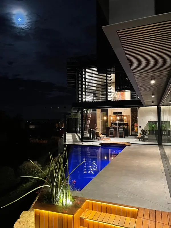 Piscina iluminada à noite, com deck de madeira com luz embutida e a casa acesa ao fundo.