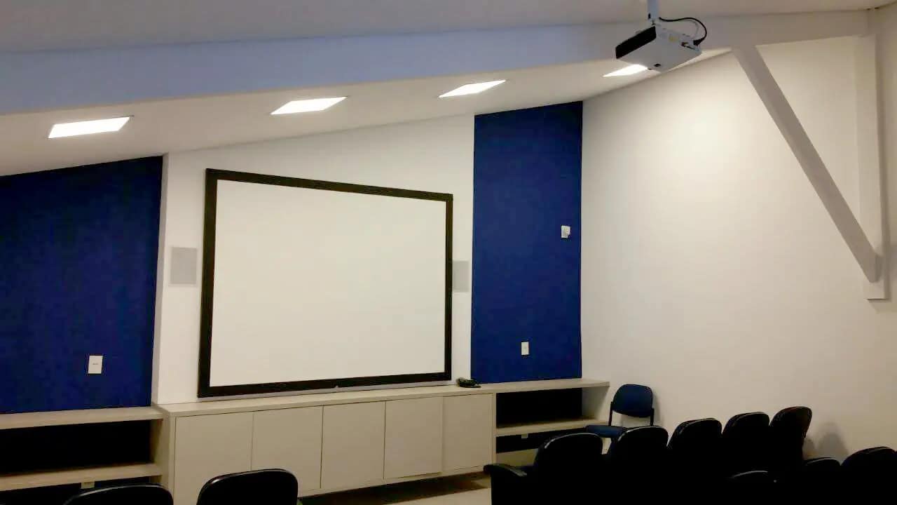 Sala de treinamento com tela de projeção fixa, projetor no teto e cadeiras em fileiras.