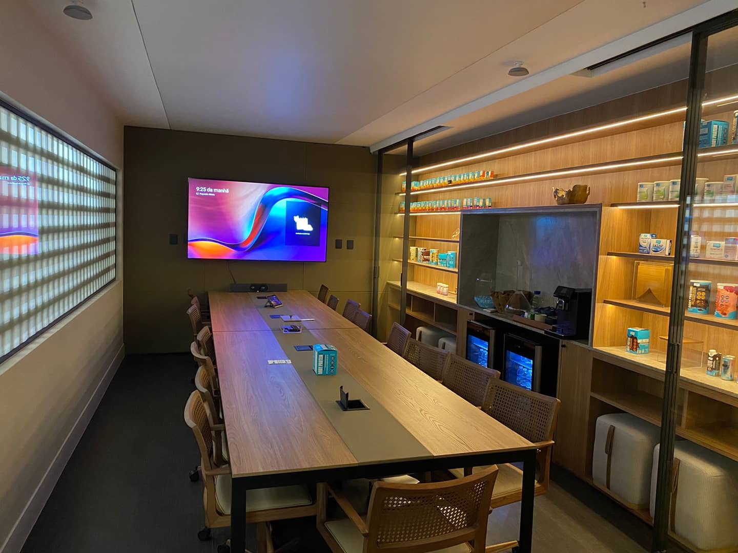 Sala de reunião com mesa longa de madeira, monitor na parede e vitrines iluminadas ao lado.