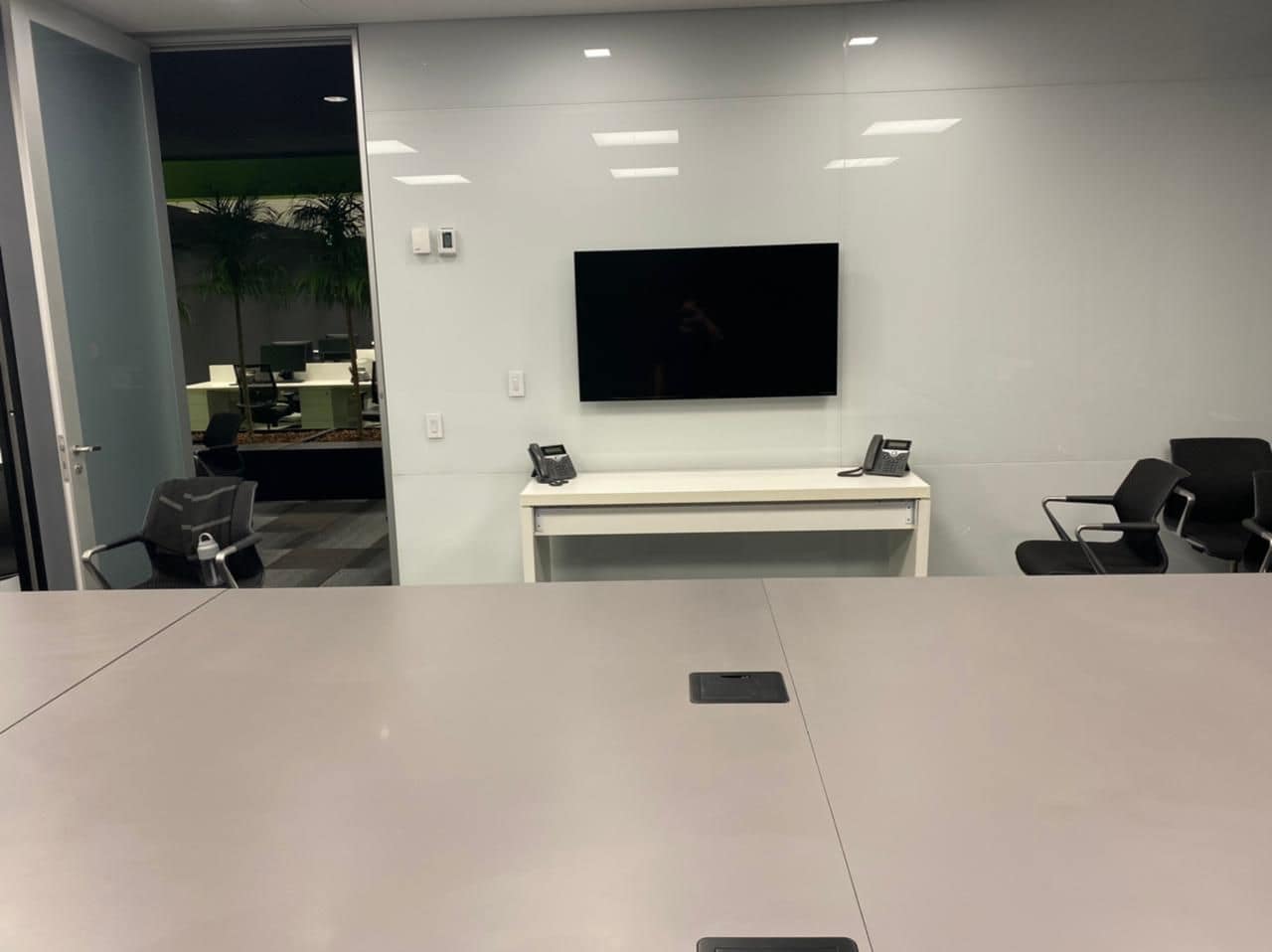 Sala de reunião com mesa branca, monitor na parede e telefones de conferência no aparador.