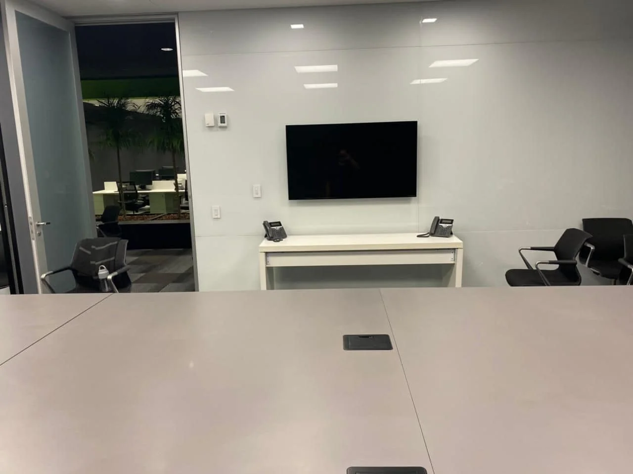 Sala de reunião com mesa branca, monitor na parede e telefones de conferência no aparador.