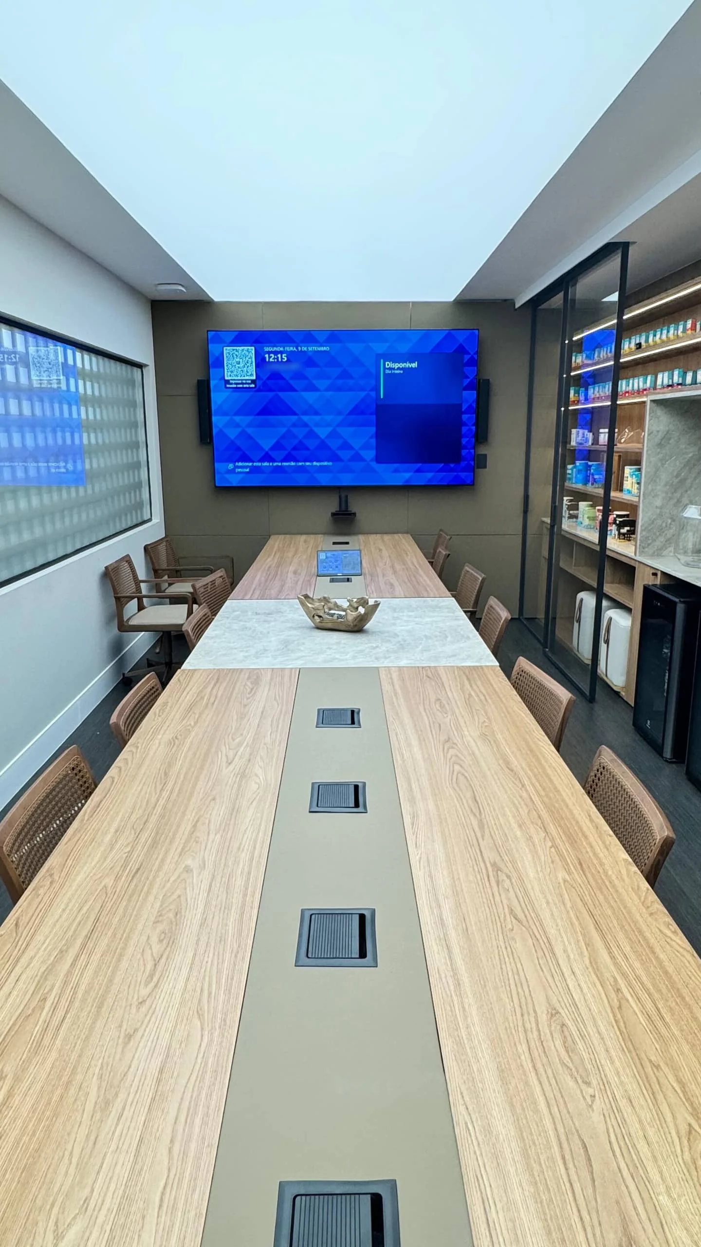 Sala de reunião com mesa clara, pontos de conexão na mesa e monitor ao fundo.