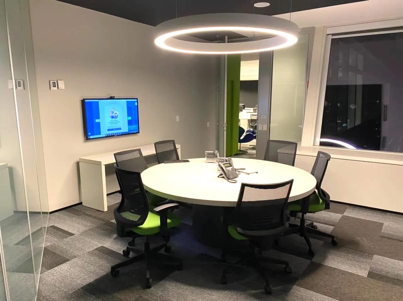 Sala de reunião com mesa redonda, anel de luz no teto e monitor na parede.