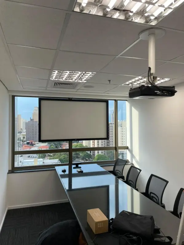 Sala de reunião com projetor no teto, tela de projeção e mesa longa com cadeiras.