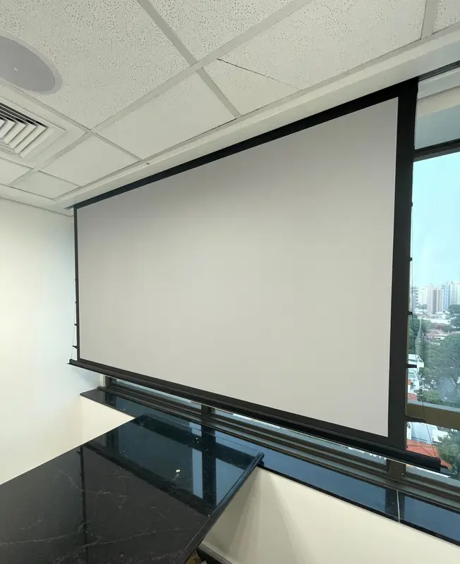 Sala de reunião com tela de projeção retrátil descida junto à janela e mesa de pedra escura.