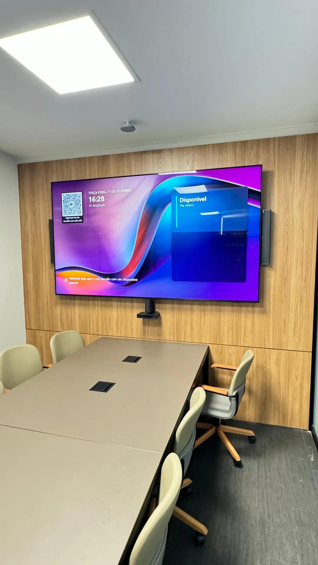 Sala de videoconferência com monitor na parede ripada e mesa com caixas de tomada.